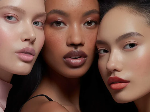 Three women with different skin tones pose closely together against a pink background, showcasing smooth complexions and natural makeup.